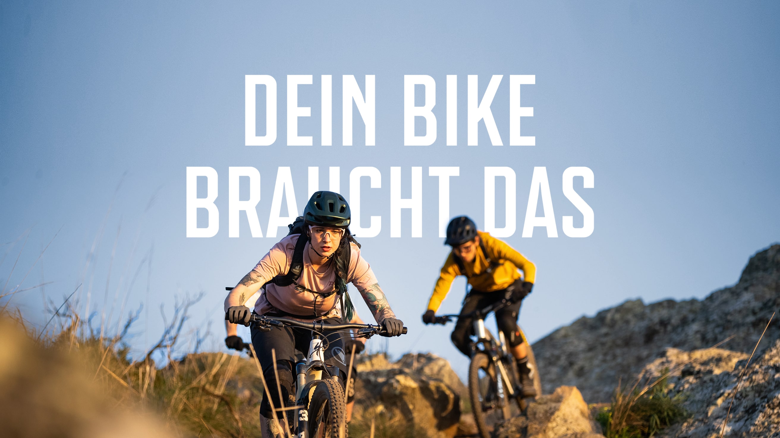Stimmungsstarkes Markenmotiv mit zwei Mountainbiker:innen auf einem felsigen Trail – mittig platziert der Claim „Dein Bike braucht das“, der die Produktrelevanz emotional auflädt.