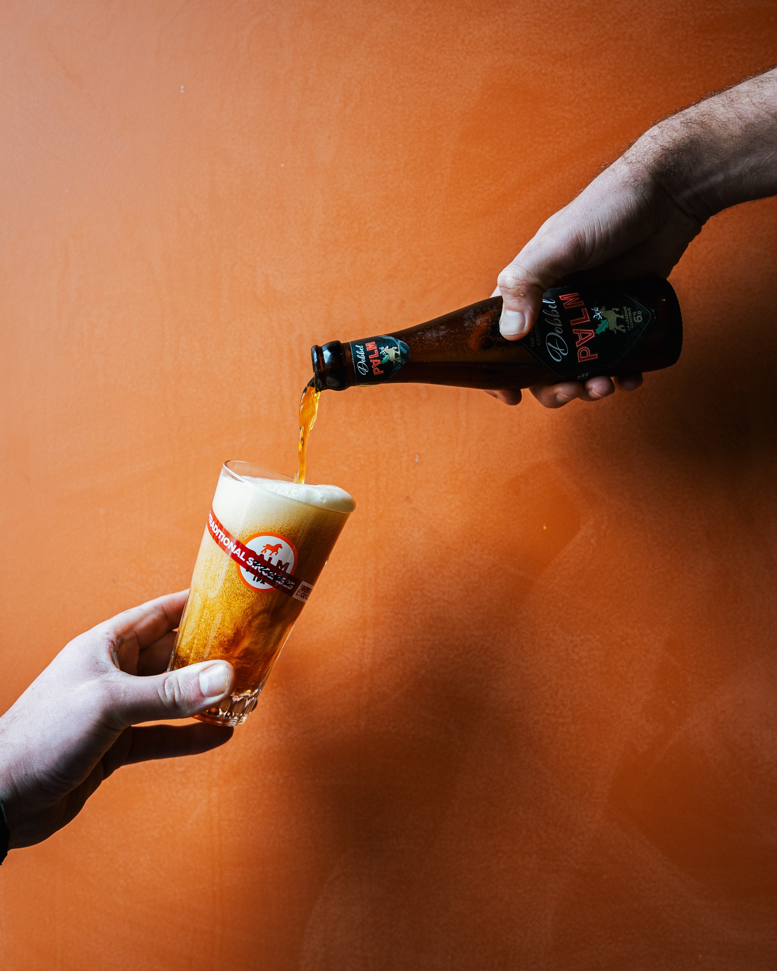 Een fles bier wordt in een glas geschonken tegen een oranje muur