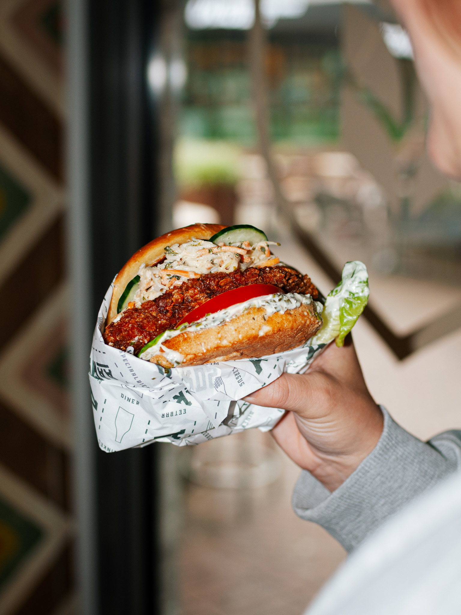 Hand houdt een burger met knapperige kip, tomaat en koolsalade in papier vast