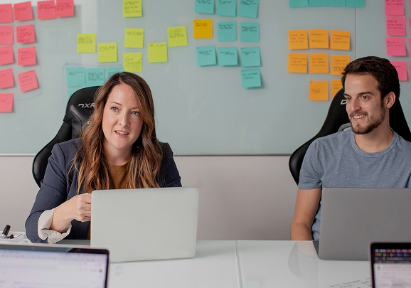 Two people sitting at a table with laptops in front of a whiteboard covered with colorful sticky notes.