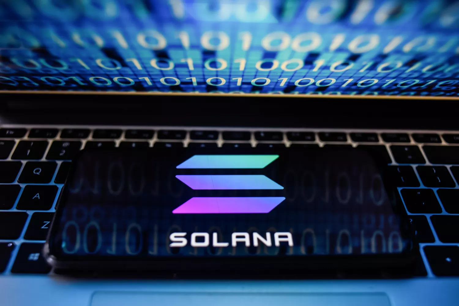 photo illustration a Solana logo seen displayed on a smartphone with binary code on a laptop screen.