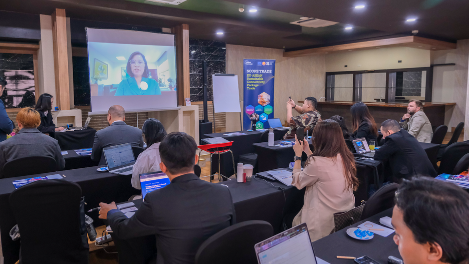 People attending a conference, watching a speaker on a screen at the front of the room.