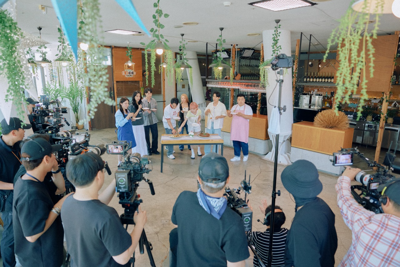 A film crew with multiple cameras surrounds the Hakka Kitchen cast around a tasting table during a shoot at a Seoul restaurant