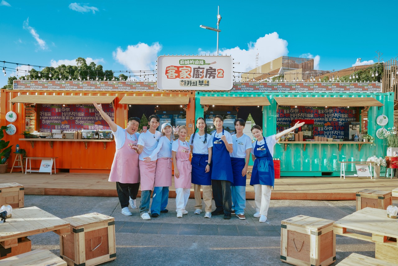 Cast members of the variety show Hakka Kitchen pose with arms raised in front of two colourful HAKKA-branded shipping-container food stalls