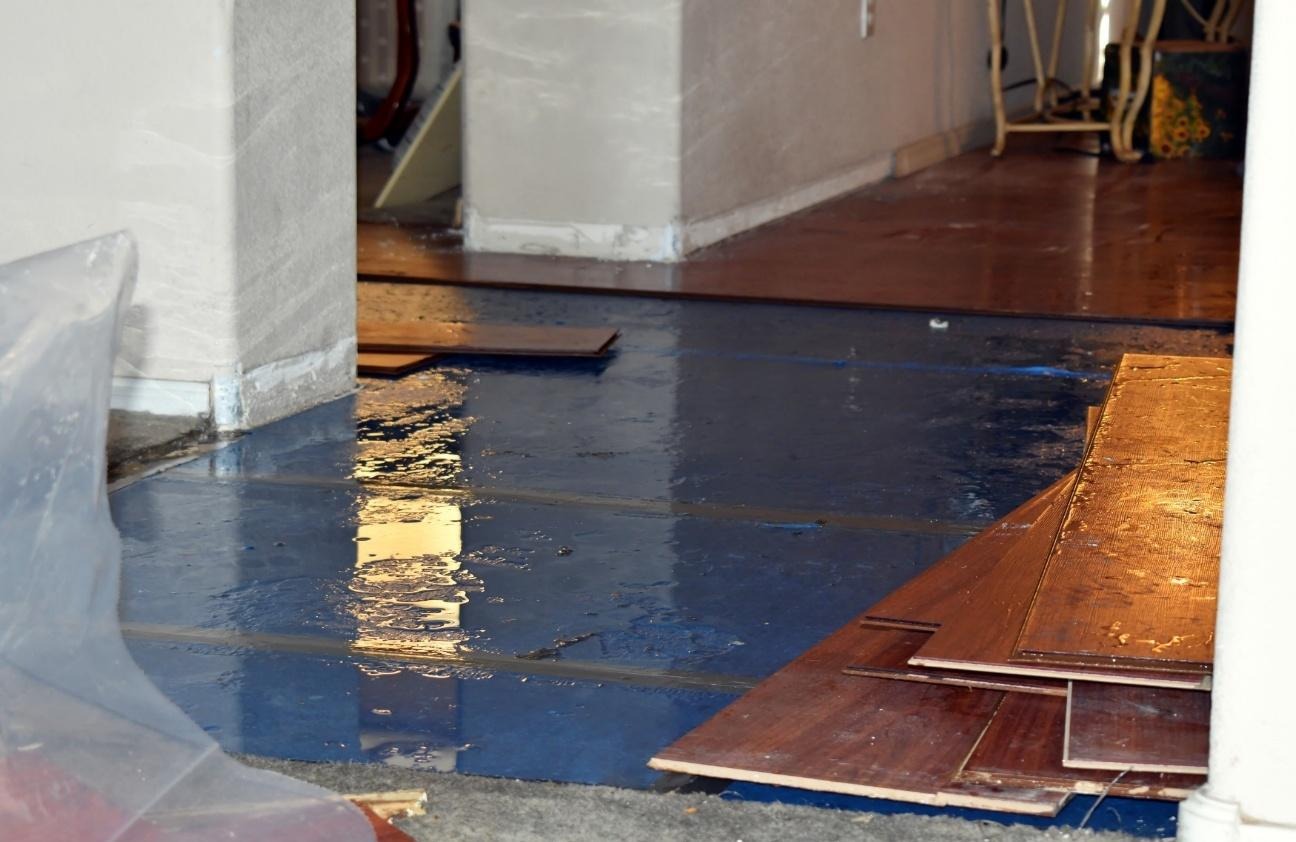The image shows a partially flooded room with exposed wooden floorboards and a blue tarp covering some furniture.AI-generated content may be incorrect.