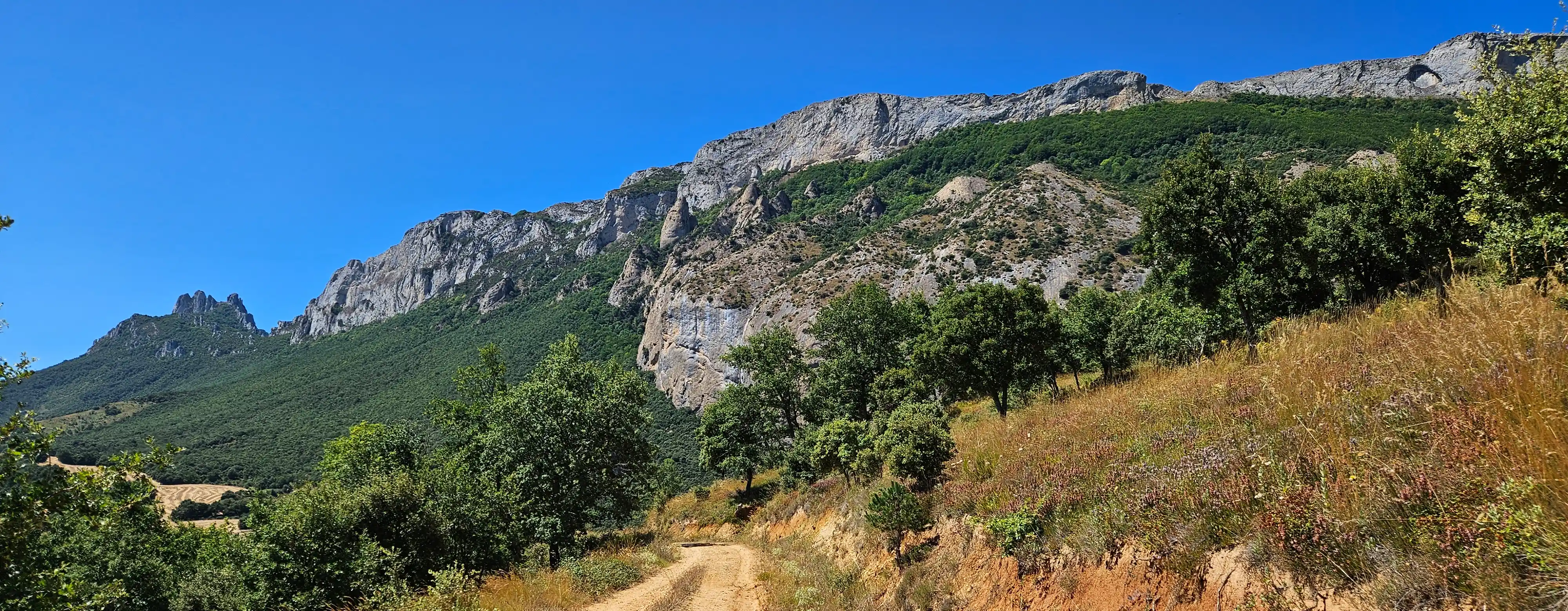 Piste en terre montant au pied d’une barre rocheuse sous un ciel bleu