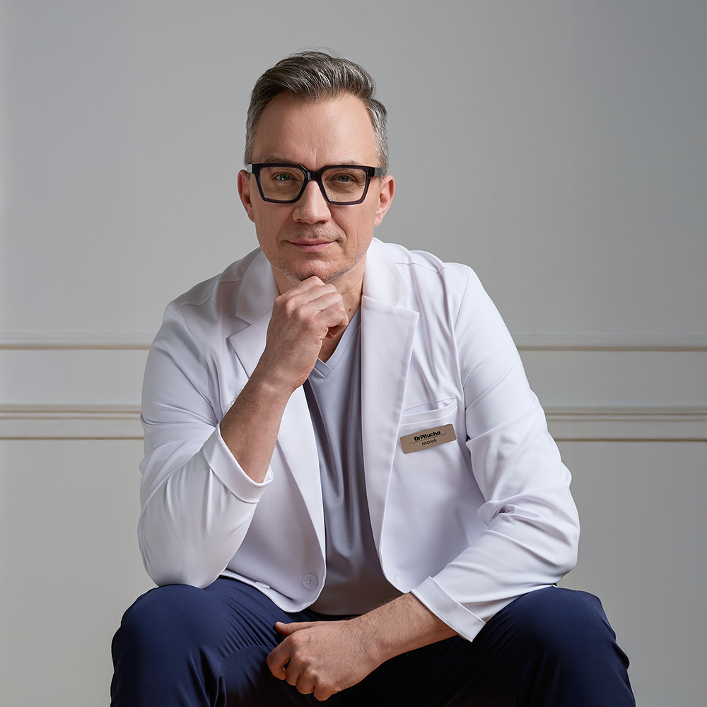 Mężczyzna w białym fartuchu lekarskim i okularach patrzy prosto, podpierając podbródek dłonią.