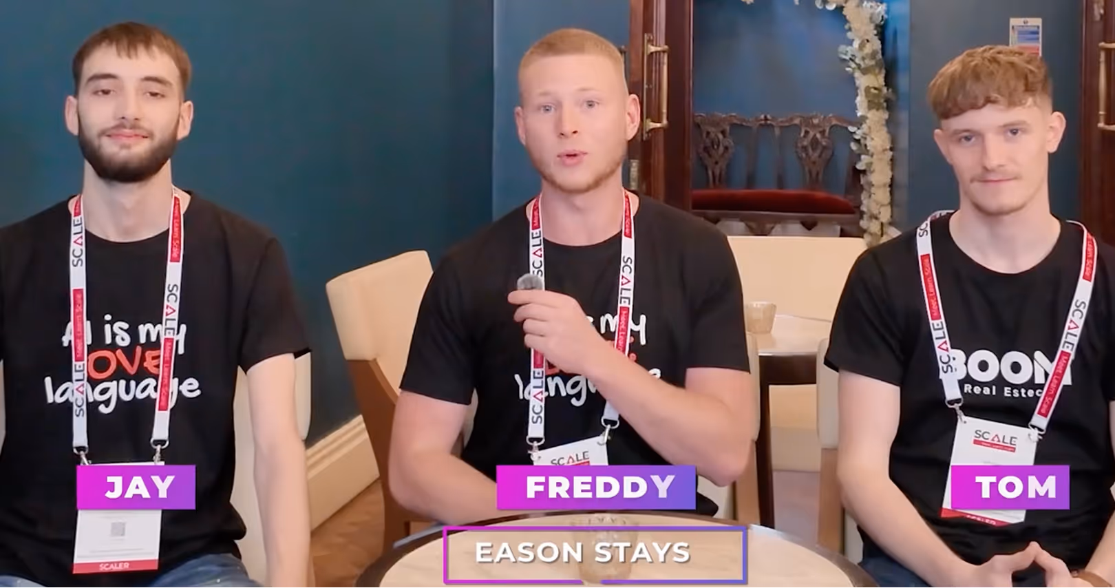 Three young men, Jay, Freddy, and Tom, sit at a round table wearing black t-shirts and conference lanyards.