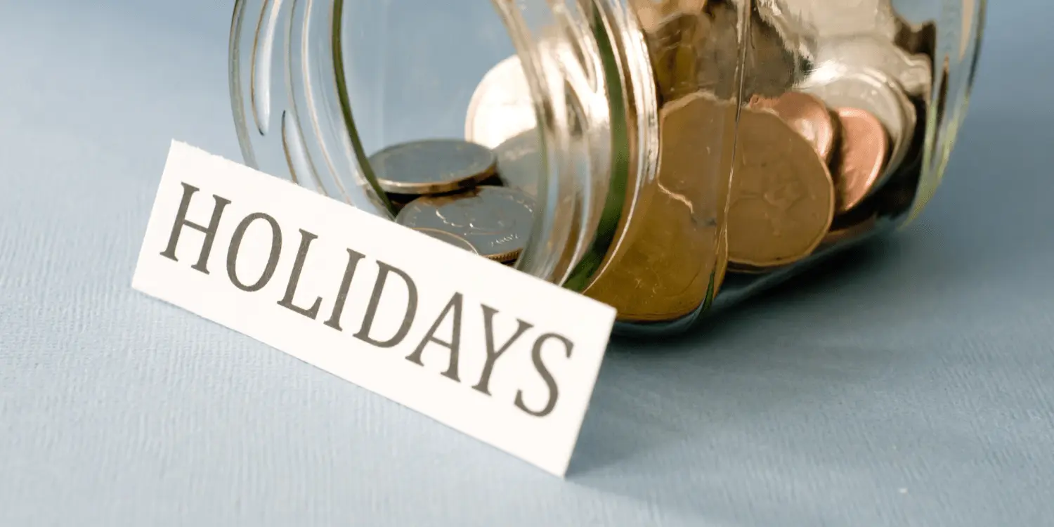 A glass jar filled with coins and a sign that says holidays encouraging the 15 day holiday challenge to save money.