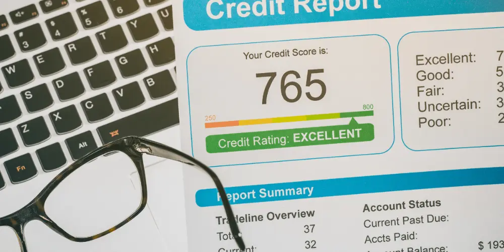 A credit report sitting on top of a laptop next to a pair of glasses with a score of 765 and report summary detailing the account history.