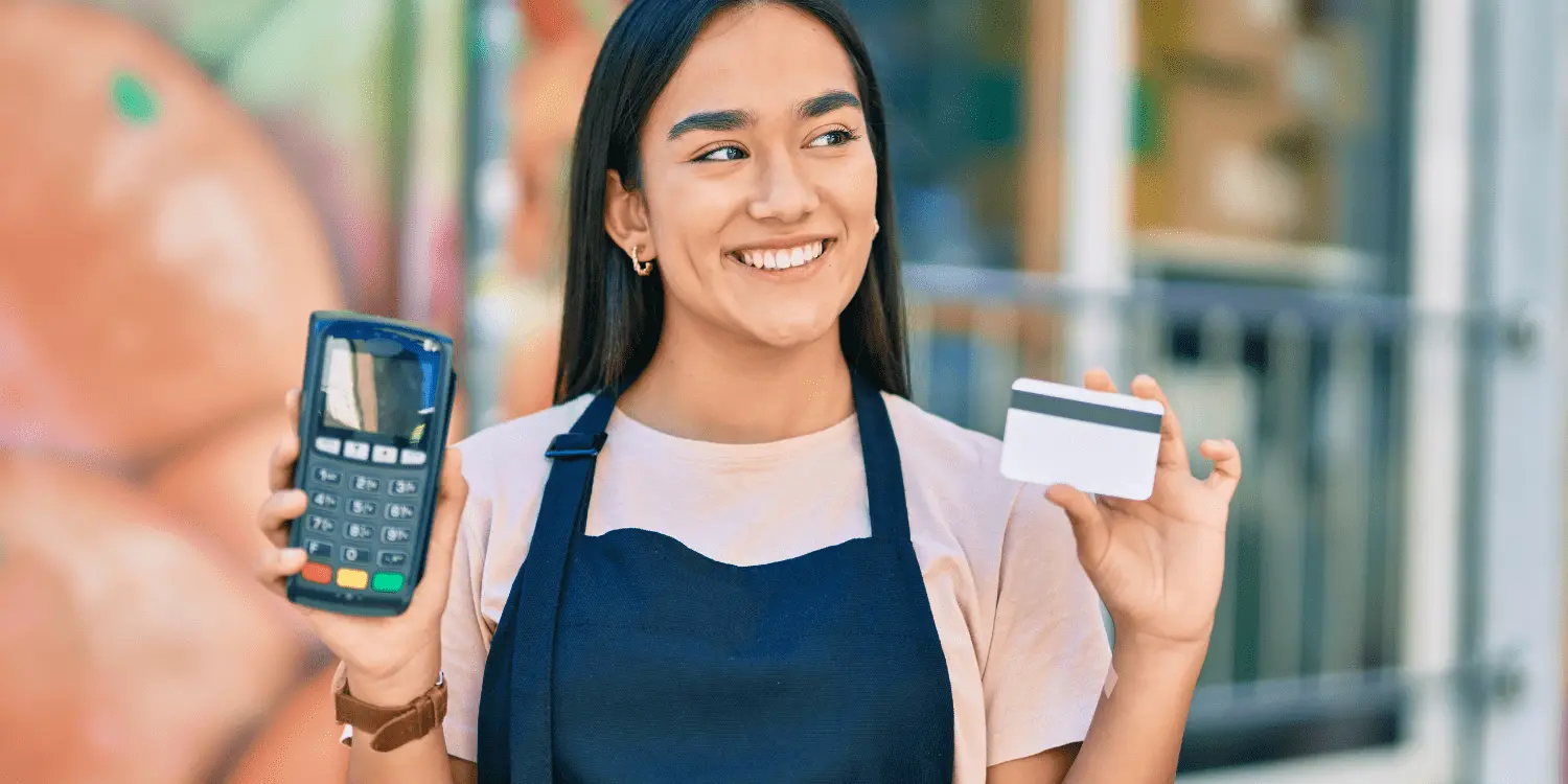 A smiling retail person holding a credit card offered by their store and a credit card reader encouraging to open credit cards with care.