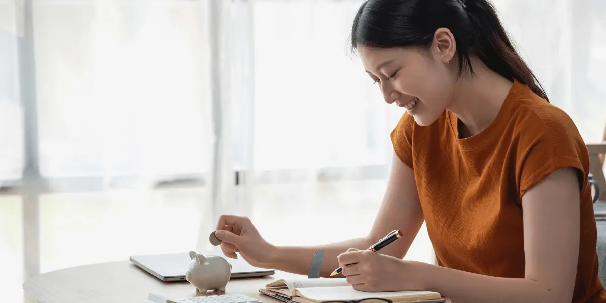 A person puts coins into a white piggy bank while writing notes in a notebook about their saving strategies mentioned in this article.