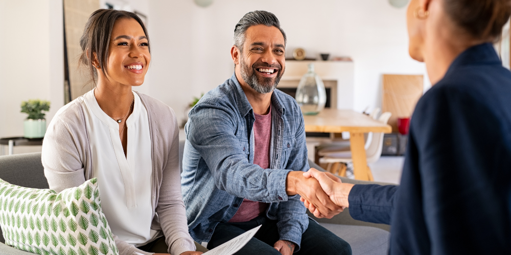 A man and woman meet with a real estate agent, shaking hands while discussing mortgage rates and optimal deals.  