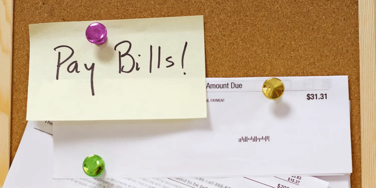 A post-it with the word "pay bills" pinned on a cork board, with billing statements pinned as well.
