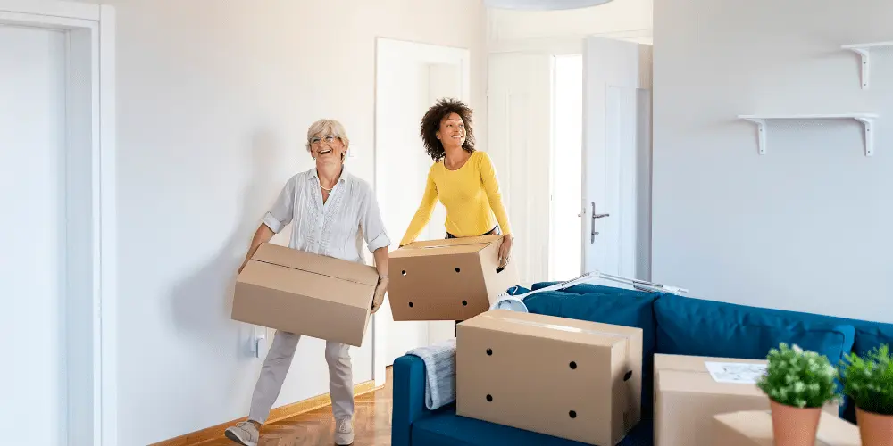 Two roommates happily moving boxes into a new apartment, showing the benefits of shared housing costs.