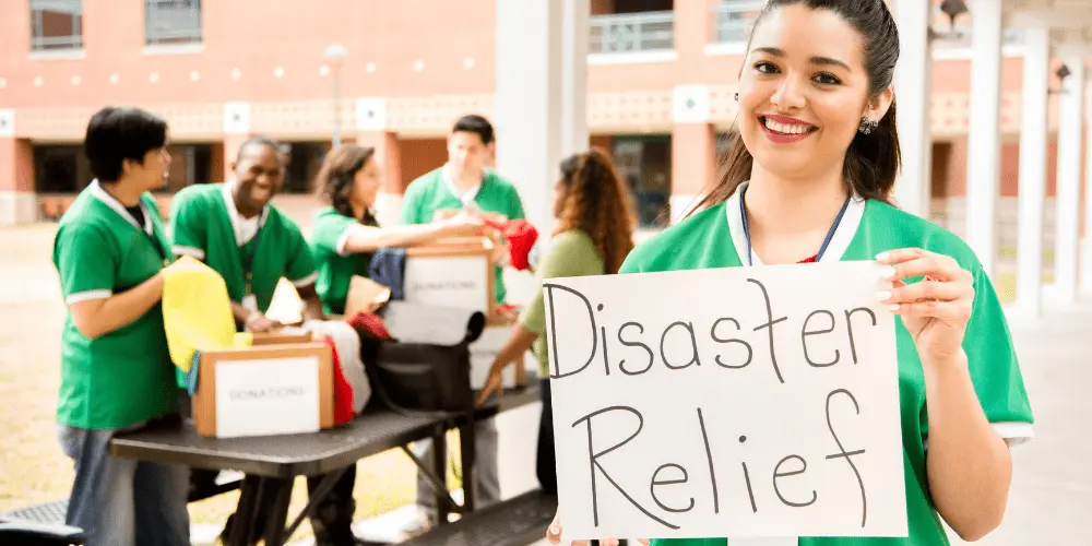 Smiling volunteer holds a Disaster Relief sign while a team distributes donations to support FEMA disaster assistance efforts.

