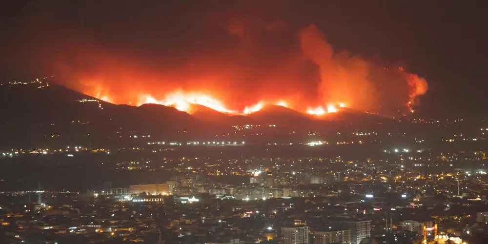 Wildfire blazing near a city at night, representing natural disasters that can threaten homes, safety, and financial stability.