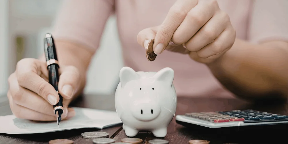 Hand dropping coin into savings jar near laptop, showing building an emergency fund for unexpected expenses