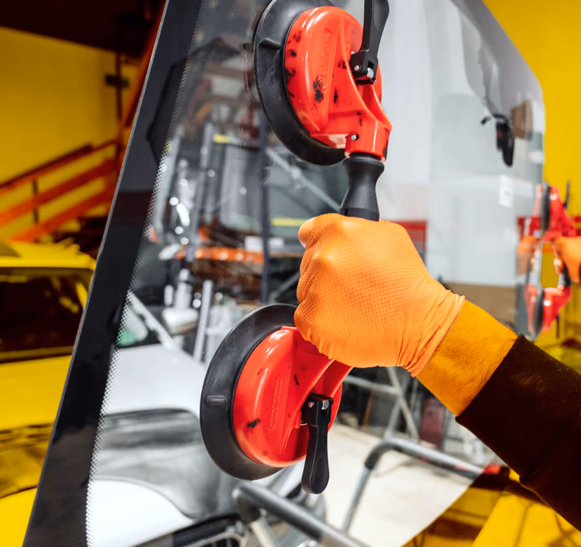 Monteur im orangen Handschuh trägt eine Windschutzscheibe mit doppeltem Saugheber durch die Werkstatt