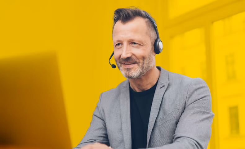 Ein freundlicher Mann im Sakko und einem Headset arbeitet am Laptop.