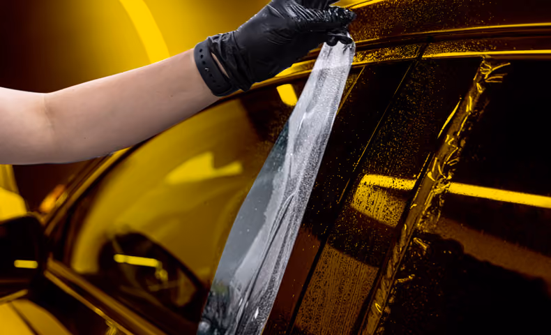 Eine Hand mit schwarzen Nitril Handschuh bringt Folie auf einer Seitenscheibe eines Autos zur Tönung auf.