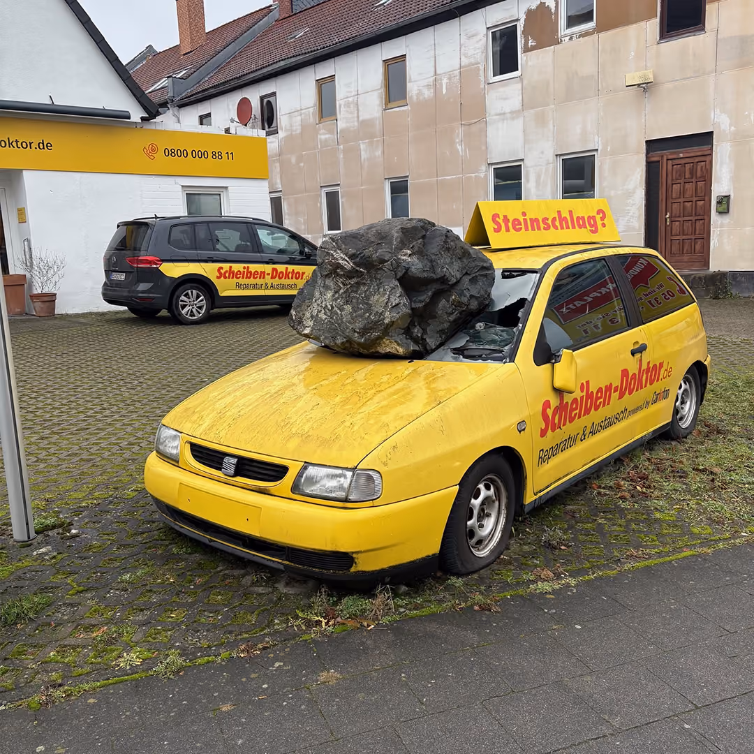 Gelbes Scheiben-Doktor-Werbeauto mit großem Felsbrocken auf der Scheibe und Schild „Steinschlag?“