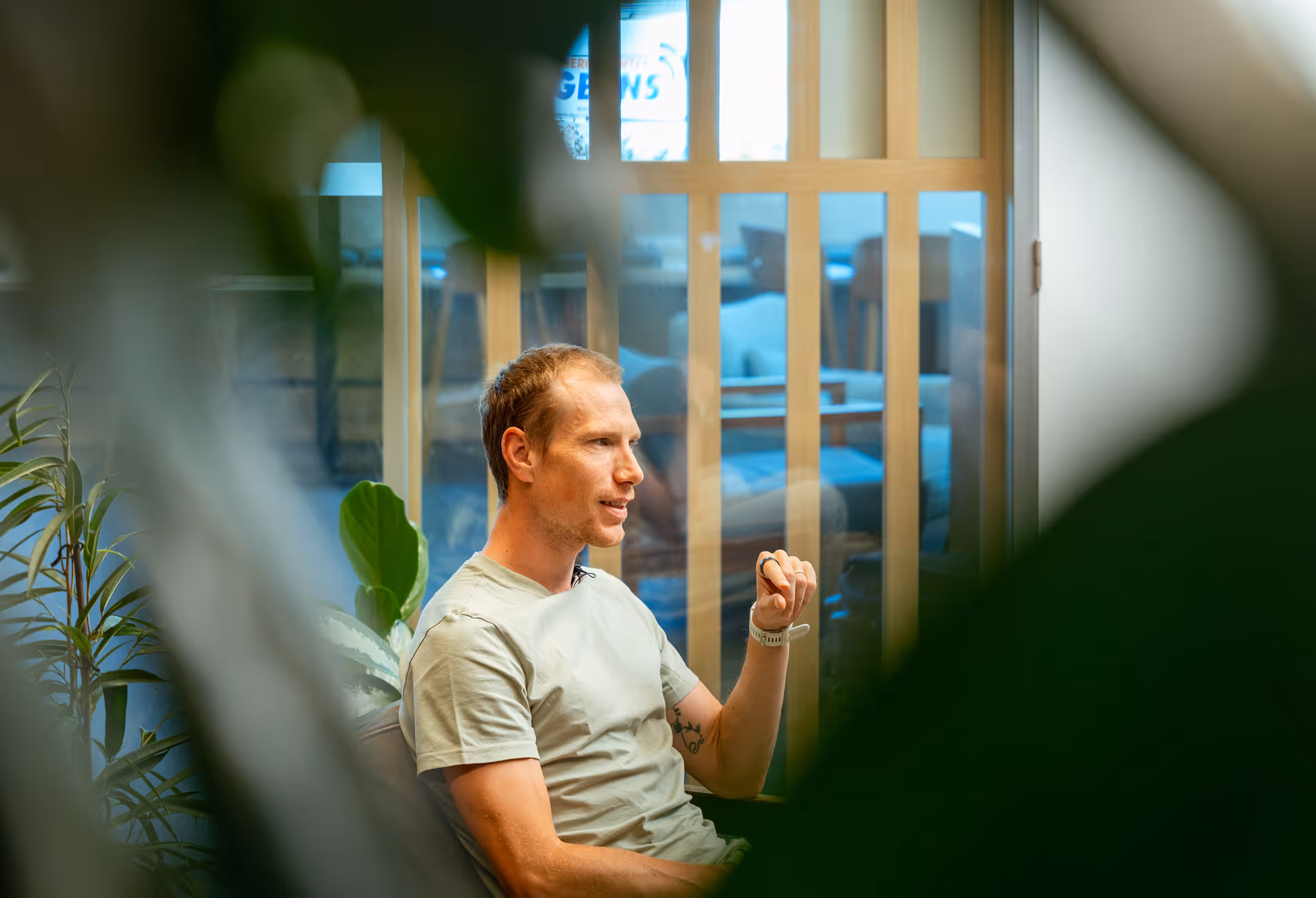 Man with light skin and short hair wearing a light shirt, sitting and gesturing while speaking in a room with glass paneling and indoor plants.