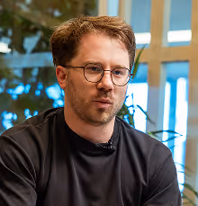 Man with glasses and short brown hair wearing a black shirt sitting indoors with plants and windows in the background.