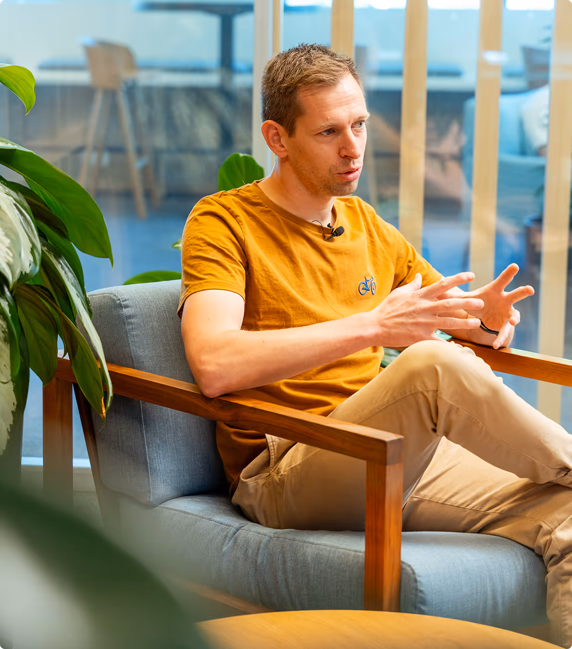 Man in a mustard yellow shirt with a bicycle logo, sitting on a gray armchair, speaking with hand gestures in a modern indoor space with plants.