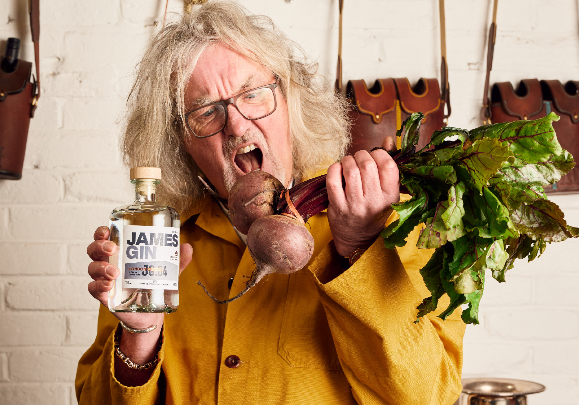 James May holding a bottle of his London Drizzle gin and going in to bite 2 beetroots