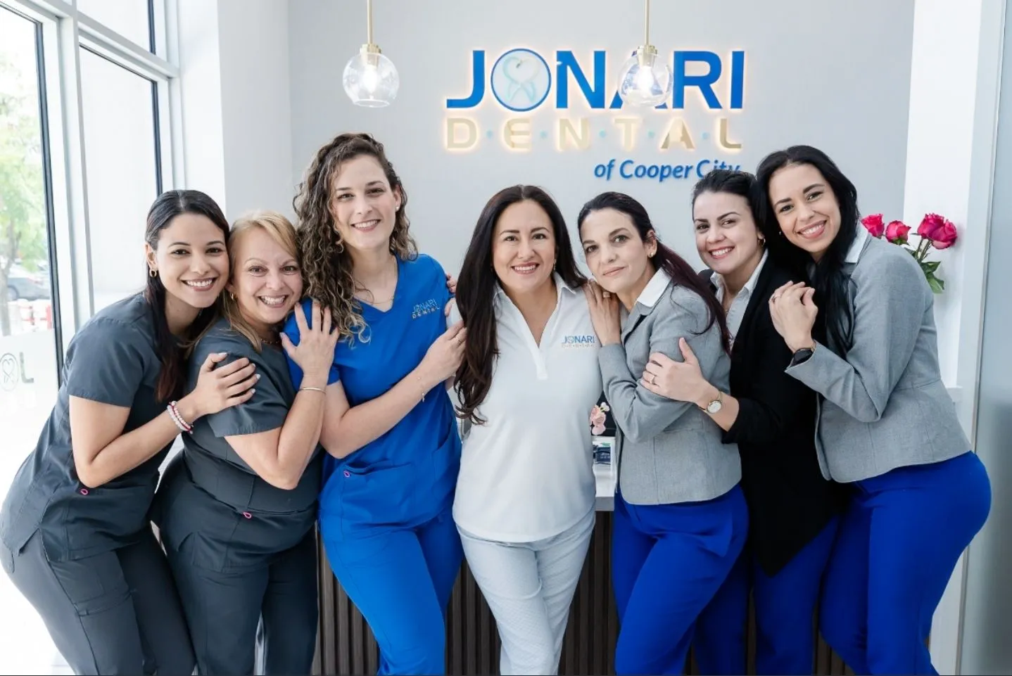 Seven smiling women standing closely together in a dental office with a Jonari Dental sign in the background.