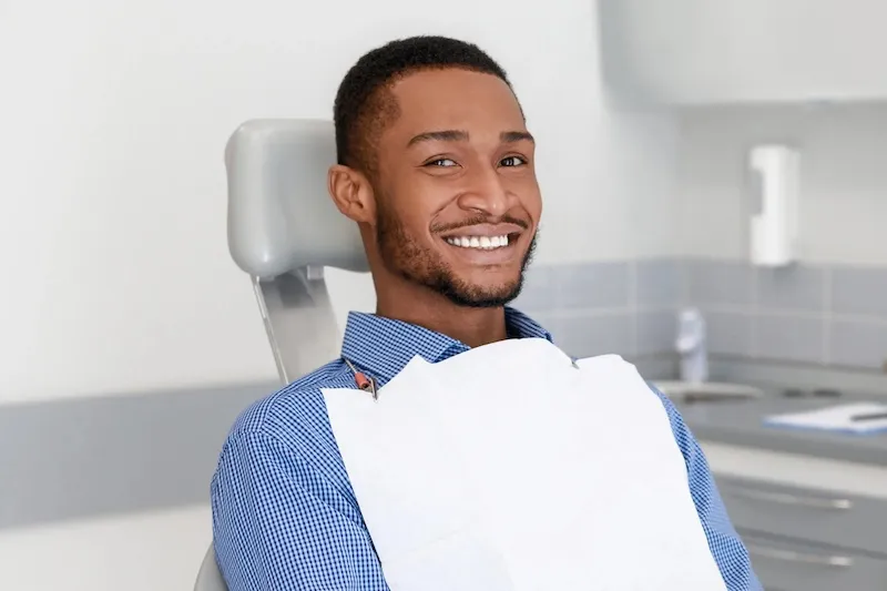 Smiling man sitting in a dental chair wearing a blue checkered shirt and a white protective bib.