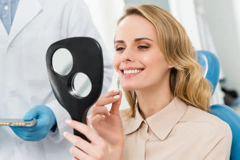 Smiling woman holding a dental shade guide to her teeth while a dentist holds additional tooth shades nearby.