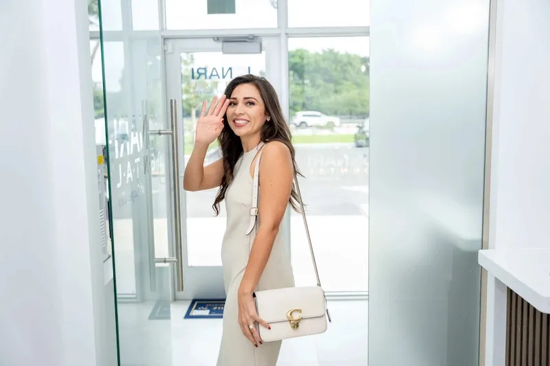 Smiling woman in a sleeveless beige dress waving goodbye indoors near a glass door.