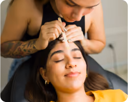 Woman receiving eyebrow grooming with a brush from a tattooed beautician.