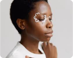 Close-up of a person with dark skin and short curly hair, eyes closed, with white cloud-like designs around their eyes.