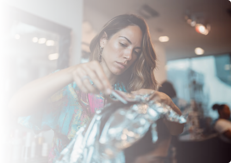 Woman with orange hair concentrating while selecting nail polish among various bottles and decorative nail art on her fingers.