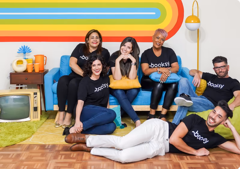 Group of six smiling diverse people wearing black Booksy shirts standing in front of a rainbow-colored wall, making heart shapes with their hands.