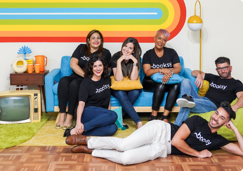Group of six smiling diverse people wearing black Booksy shirts standing in front of a rainbow-colored wall, making heart shapes with their hands.