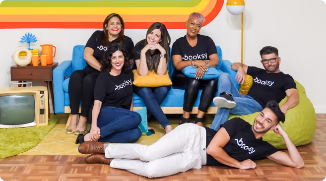Six diverse people wearing black Booksy t-shirts standing in front of a rainbow-colored wall, making heart shapes with their hands.