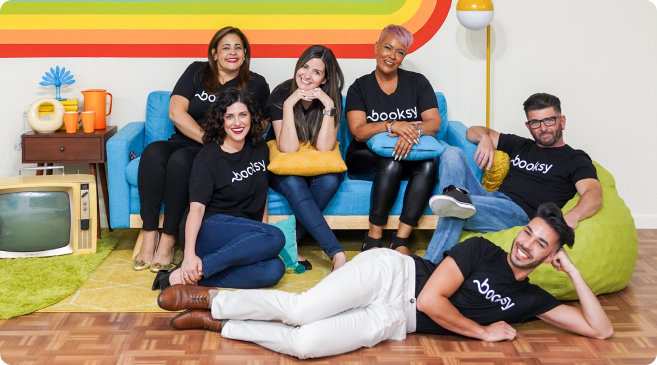 Six diverse people wearing black Booksy t-shirts standing in front of a rainbow-colored wall, making heart shapes with their hands.