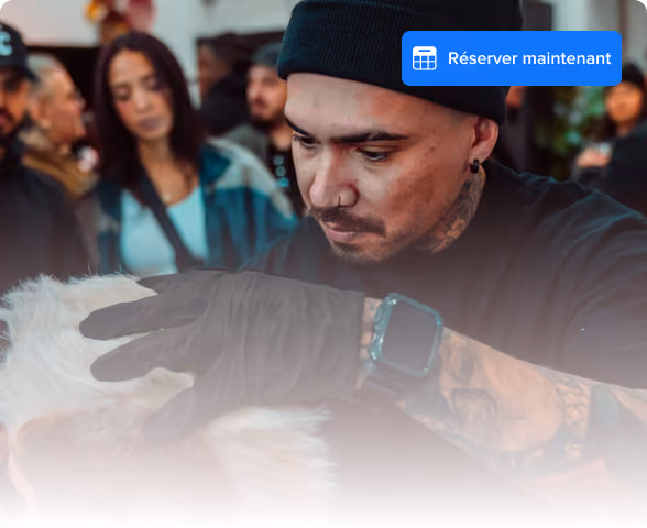 Homme tatoué portant un bonnet noir et des gants toilettant un chien blanc à l'intérieur avec des gens en arrière-plan.