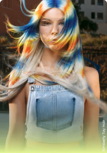 Young woman with long hair dyed in vibrant blue, yellow, and orange colors, wearing a denim overall top.