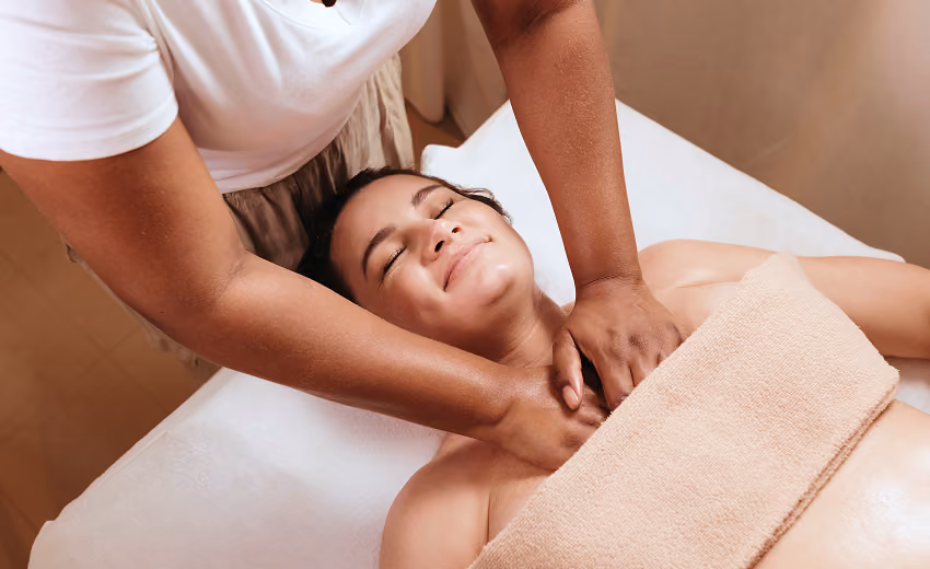 Woman with eyes closed receiving a relaxing head massage while lying on a towel with a lit candle in the background.