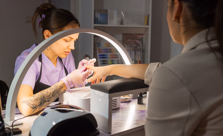 Manicurist wearing glasses and an apron files nails of a seated woman with blonde hair in a nail salon.