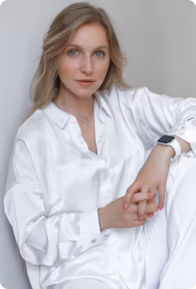 Femme blonde aux vagues lâches, vêtue d'un chemisier et d'un pantalon en satin blanc, assise, les mains jointes, regardant la caméra.