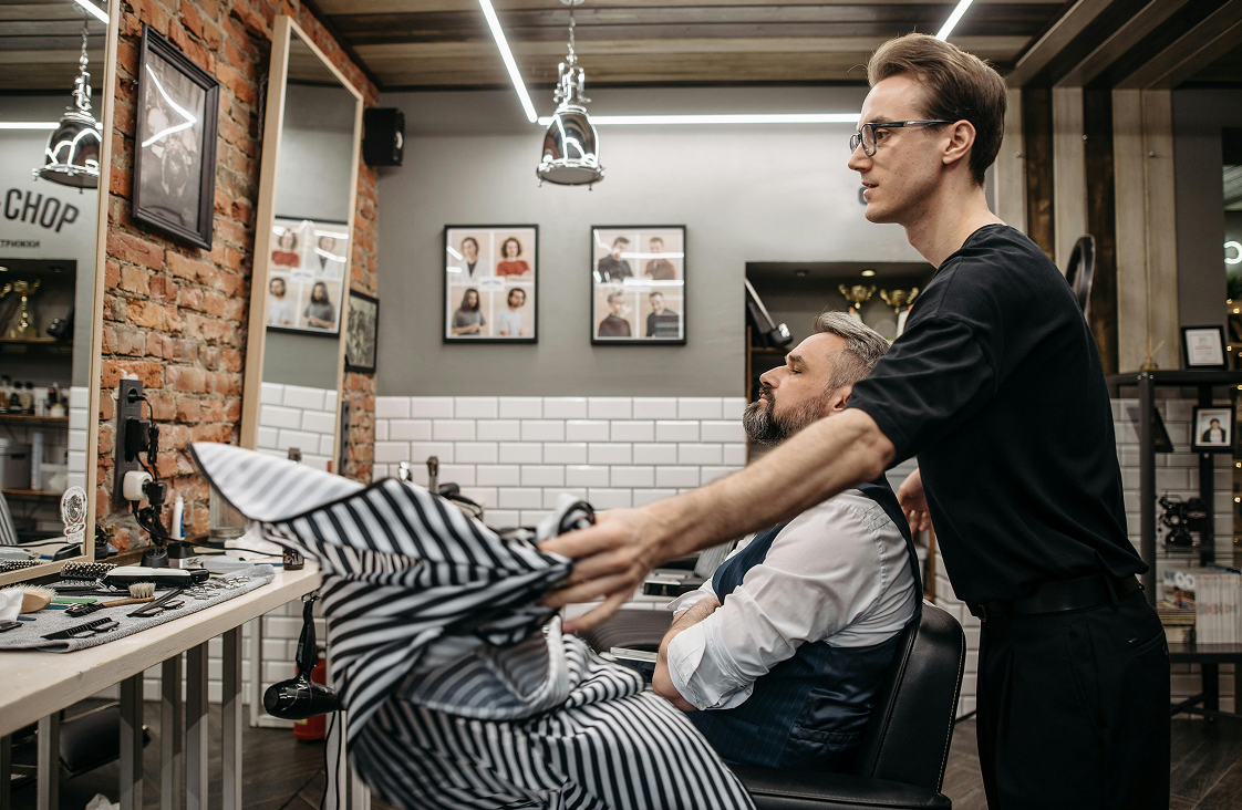 Barber puts on striped cape onto male client with beard.