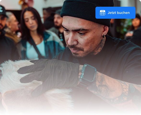 Tattooed man wearing a black beanie and gloves grooming a light-colored dog indoors with people in the background.