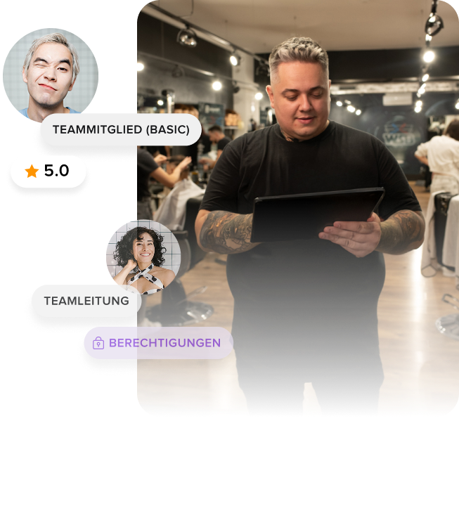 Man with tattoos wearing a black shirt using a tablet in a busy barber shop with blurred background of customers and stylists.