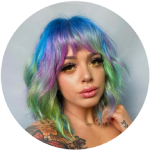 Woman with multicolored blue, green, and purple hair and tattoos on shoulder posing against gray background.